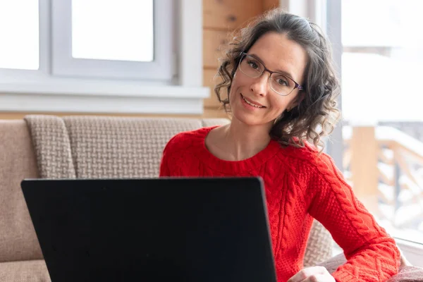 A woman in a red jacket works and watches social networks on her computer. Female with a laptop is sitting on the sofa by the window.