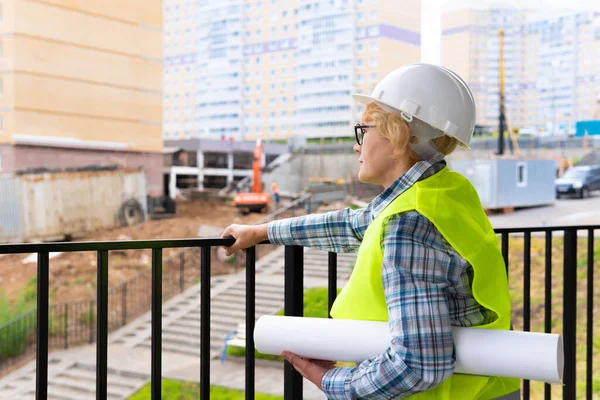 A female construction engineer is on a construction site. A middle-aged woman holds the project in her hands. She looks at the house.