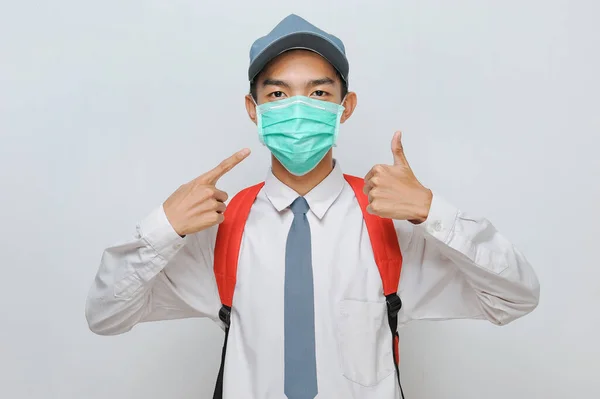 Indonesian Senior Student wearing uniform and protection face mask against coronavirus right hand pointing at face mask on his face and left hand doing okay sign, isolated on grey background