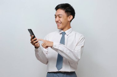 Lise üniforması giyen mutlu Endonezyalı öğrencinin portresi gri arka planda izole bir şekilde telefon ekranına bakıyor.