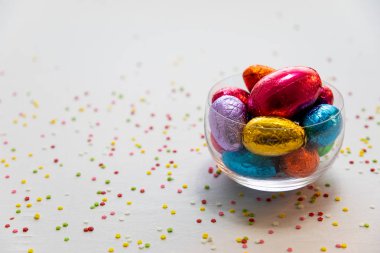 Beyaz arka plan ve bulanık konfeti ile şeffaf bir kapta renkli çikolata paskalya yumurtaları