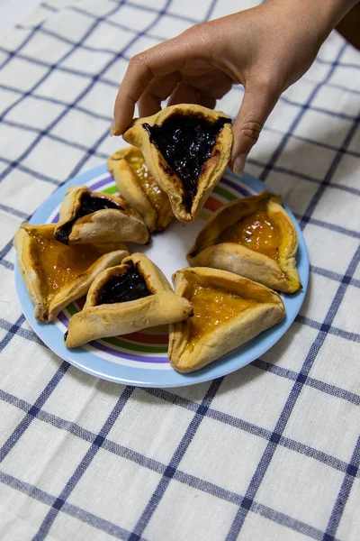 Mão mulher segurando Hamantash Purim Blueberry e geléia de damasco biscoitos na placa colorida na toalha de mesa azul e branco — Fotografia de Stock