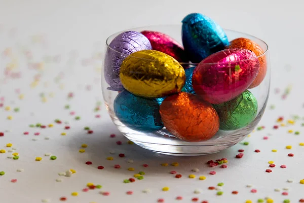 Colorful chocolate easter eggs in a transparent bowl with white background and blurred confetti — Stock Photo, Image