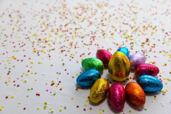 Many aligned colored chocolate easter eggs on white background and colorful confetti — Stock Photo, Image