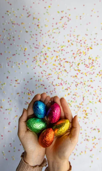 Beyaz arka planı ve renkli bulanık konfetileri olan renkli çikolatalı paskalya yumurtalarını tutan kadın elleri