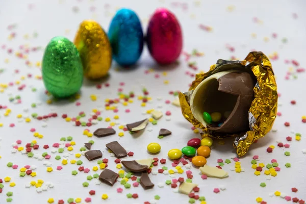 Rompere uova di Pasqua di cioccolato dorato con cioccolatini colorati all'interno su sfondo bianco con confetti sfocati colorati e altre uova colorate — Foto Stock