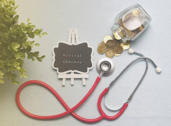 Top view of medical stethoscope, jar with coins and frame with medical checkup lettering on white stone background