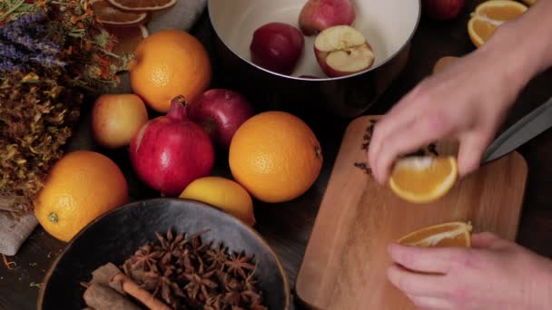 Preparación de vino caliente. Las manos femeninas cortan naranjas y limones en una tabla de cortar de madera y los ponen en una cacerola negra. Limones frescos, naranjas, granadas y hierbas secas en el fondo — Vídeos de Stock