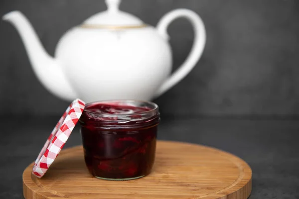 Cherry jam, jam, jelly in a glass jar with a red and white lid next to a wooden round board, board. Against the background of a white teapot. Gray background. Place for text — Stock Photo, Image