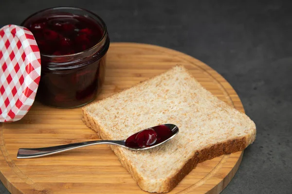 Cherry jam with berries in a glass jar with an open red and white lid next. Next to a wholegrain toast with an empty teaspoon. On a wooden board. Gray background — Stock Photo, Image