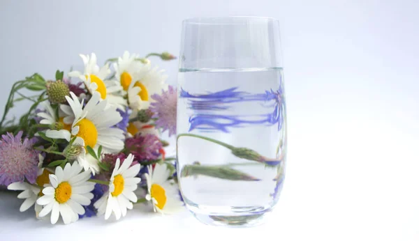 Ein Strauß Wildblumen liegt neben einem Glas Trinkwasser. Ein Strauß Gänseblümchen, Kleeblumen, roter Mohn und blauer Kornblumen neben einer Glasschale mit Wasser. Blumen spiegeln sich im Glas — Stockfoto
