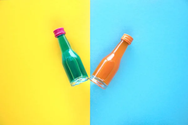 Two small glass bottles with a blue and orange cocktail on a blue and yellow background. Two multi-colored small bottles with multi-colored covers.