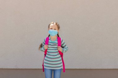 Girl wearing a protective mask with a backpack behind his back in the schoolyard on the first school day after isolation or vacation. Children are happy to go back to school clipart