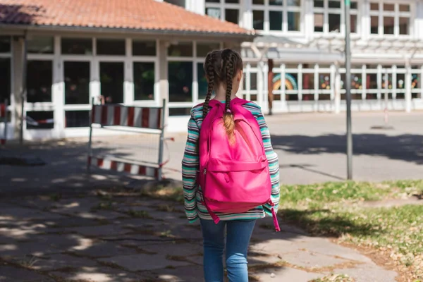 A girl with a pink satchel on her back approaches the school door. The first day of school, the children are ready to study after the holidays. Education, lessons after the pandemic.