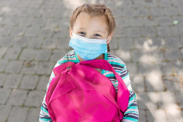 Yüzünde koruyucu bir tıbbi maske olan pembe sırt çantalı okul çağındaki bir kızın yakın plan portresi. 2020 'de yeni okul yılı. Eğitim kurumlarında çocukların korunması.
