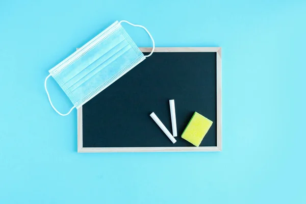 Graphite school board with a piece of chalk, a sponge and a medical protective mask on a blue background. Copy space. The beginning of the new school year. Virus protection in educational institutions