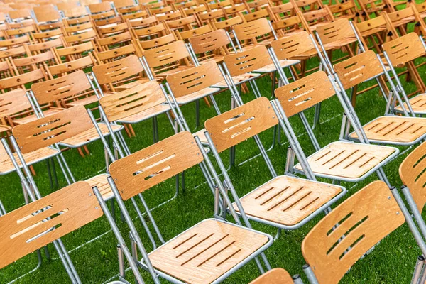 Lots of wooden chairs stand outside in the park in the rain. Empty auditorium, green grass, trees and chairs with water drops.