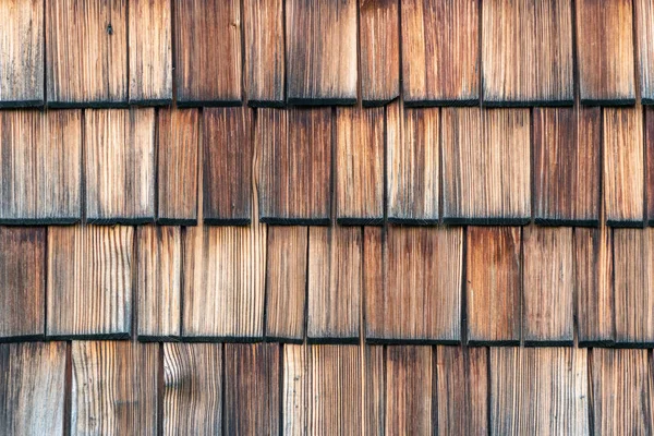 Weathered and faded from the sun wooden shingles that are the facade of an alpine house. Wooden texture in shades of gray and orange. — Stock Photo, Image