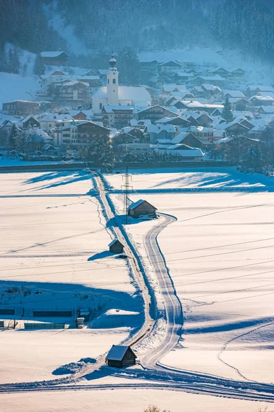 Ευθεία, χιονισμένο δρόμο χειμώνα προς Haus, μικρή πόλη στο Schladming Dachstein. Στο παρασκήνιο ο πύργος εκκλησία πάνω από την πόλη και Άλπεις βουνά — Φωτογραφία Αρχείου