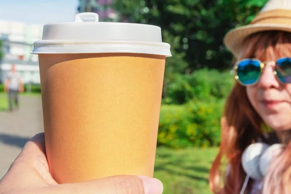 Street cup of coffee in the summer. A cup of coffee is shown in close-up. There is a place for text. In the background, a girl with glasses and a hat looks at a cup of coffee