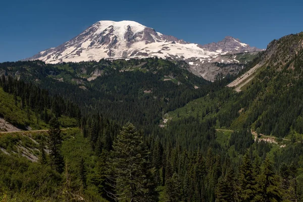 Uma montanha coberta de neve, o Monte Rainier, na primavera, com uma floresta de pinheiros verdes exuberantes em primeiro plano e estradas varrendo a imagem — Fotografia de Stock