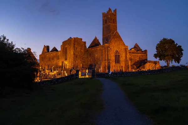 The ruins of the medieval Quinn Abbey in Quinn, Ireland lit up against a deep blue sunset sky, shot looking down the footpath leading to the abbey nobody in the image
