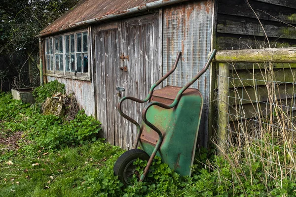 Заброшенный сарай с разбитым окном, зеленая ржавая тачка, прислоненная к сараю — стоковое фото