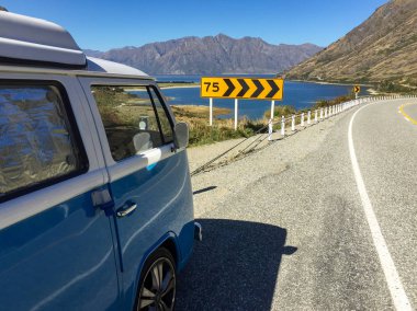 Bir vintage mavi ve beyaz campervan güzel Hawea Gölü yakınında park, keskin bir viraj ve ön planda bir hız uyarı işareti ile, açık mavi gökyüzüne karşı