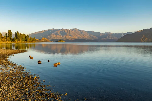 A low-level view along the edge of Lake Wanaka early in the morning as the sun rises on the hills in the distance, taken from the water\'s edge, rocks in the foreground