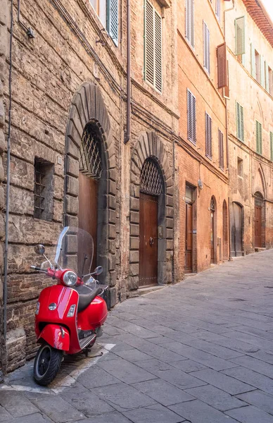 Un scooter de color rojo brillante está estacionado contra una pared en una tranquila calle lateral de Siena, la calle se curva en la distancia —  Fotos de Stock