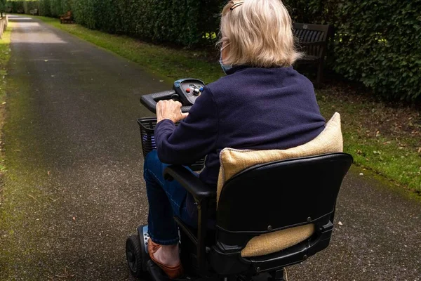 An elderly lady in a blue coat wearing a mask becasue of covid-19 enjoying the freedom of an electric mobility scooter on a quiet road.