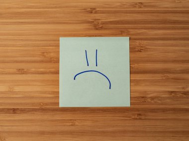 Üzücü bir ifadenin sembolünü tasvir eden bir etiket. Tahta bir yüzeye yapıştırılmış kağıt yapıştırması. Yapıştırıcı kağıt çarşaf. Tahtadaki bir kağıt parçası. Eylem için hatırlatma. Notlar için kağıt sayfa.