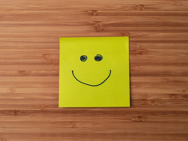 Sticker depicting the happy smiley character. Paper sticker pasted on a wooden surface. Self-adhesive paper sheet. A piece of paper on a wooden board. Reminder for action. Paper sheet for notes.