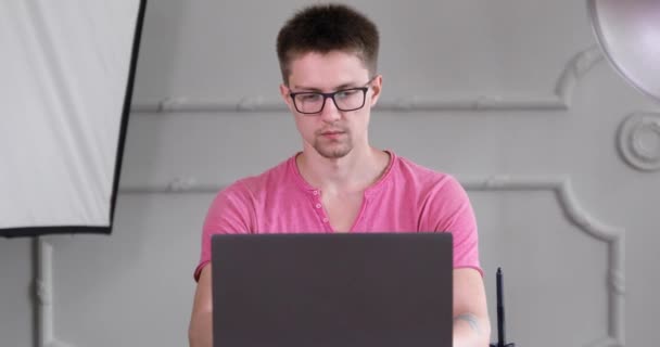 안경과 분홍색 셔츠를 남자가 테이블에 스튜디오의 노트북에서 일합니다 — 비디오