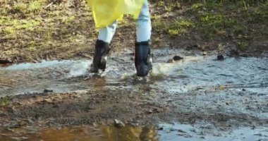 Sarı yağmurluklu ve lastik çizmeli bir kadın su birikintilerinde zıplıyor.