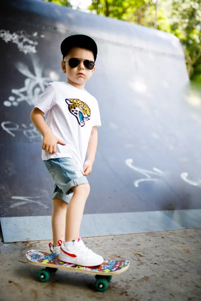 Rapaz com um skate num parque de skate. Um rapaz com óculos aprende a patinar . — Fotografia de Stock