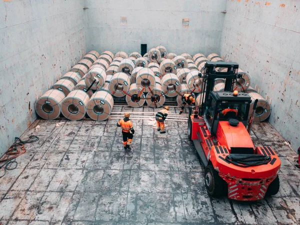 Kıyıdan kargo gemisine çelik bobinler yükleniyor. Stevedor'lar kargo ambarında çalışıyor. Hareket etmek için kaldırıcı kullanın