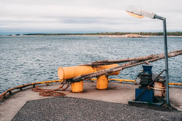 Close-up of yellow metallic mooring column or bollard on the edge of stone quay blue water background. Marine Bollard column with eye-splice on the end of rope. Loop in the end of a rope.