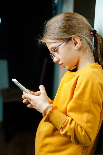 Uma garota com uma camisola amarela está segurando um smartphone em suas mãos. O problema do uso excessivo de smartphones por crianças Imagem De Stock