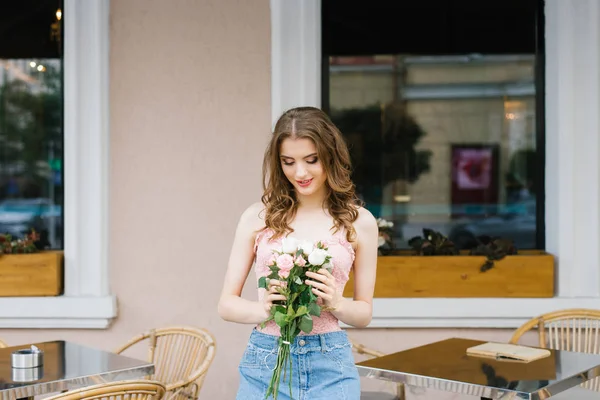 A beautiful girl in a denim skirt and a pink top is holding a bouquet of roses, looking at a bouquet and standing near an outdoor cafe chair