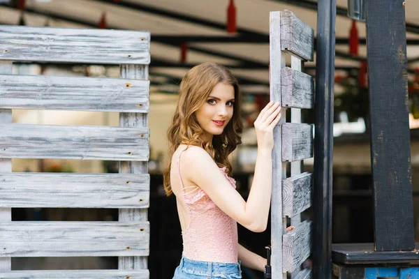 ピンクのトップと青いスカートで流れる髪を持つ美しい若い女の子は 木製の古い門の近くに立って ゲートに手を握り 夏の日 — ストック写真