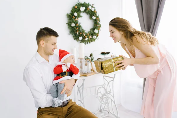 Dad holds the baby's son in a Santa Claus suit in his arms, mom gives the child a gift on the background of the Christmas tree in the living room. Christmas holiday in the family. Happiness and people