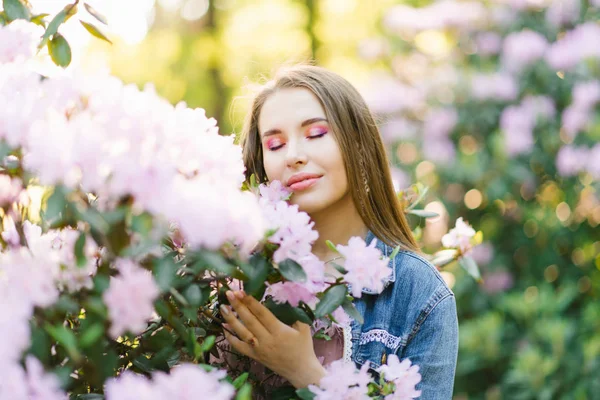 portrait of a beautiful girl with loose hair and makeup with her eyes closed near a Bush with pink rhododendron flowers in spring. The concept of peace, tranquility, happiness and enjoyment of nature