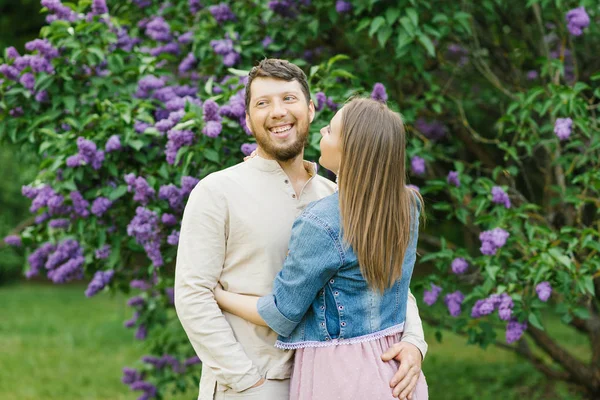 the guy hugs a beautiful girl in a pink dress and denim jacket on a background of pink flowers of lilac in the spring. the guy smiles with a wide smile. happy couple walking in spring park