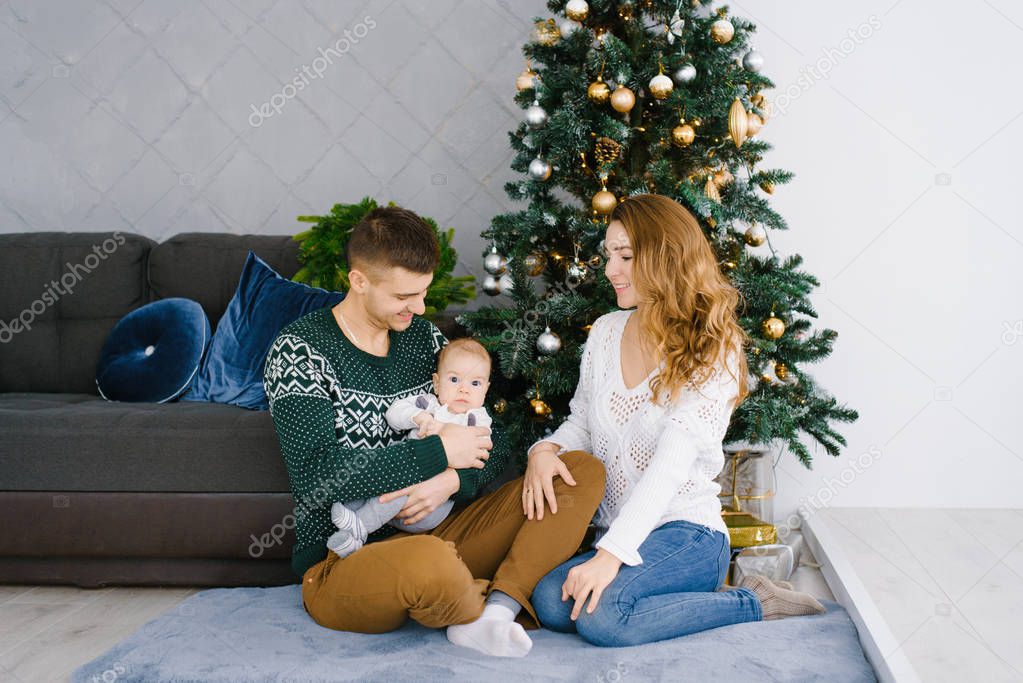 Portrait of a happy smiling and joyful family in the living room, decorated for Christmas. Father holding child in her arms, the mother looks at them and smiles. Family sitting on the floor. The concept of comfort celebrating the New Year at the Chri