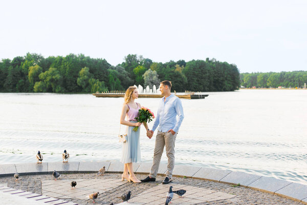 a loving couple a guy and a girl hold hands and stand on the Bank of a river or lake in a city Park. first date. girl holding a bouquet of roses