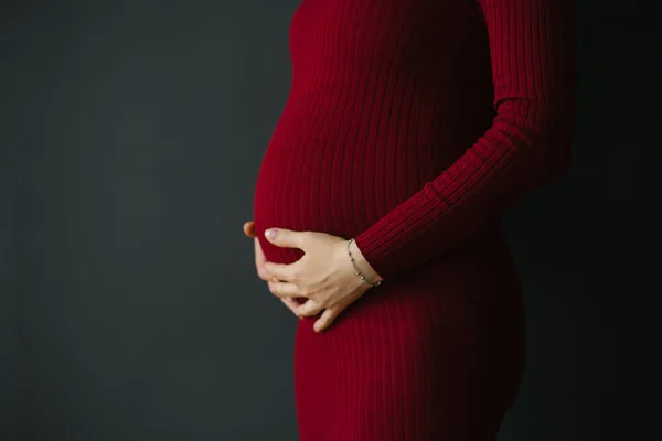ブルゴーニュのドレスを着た妊娠中の女性は 濃い灰色の背景に彼女の胃に彼女の手を保持しています 準備と期待の概念 クローズアップ 妊娠腹のクローズアップ コピースペース — ストック写真