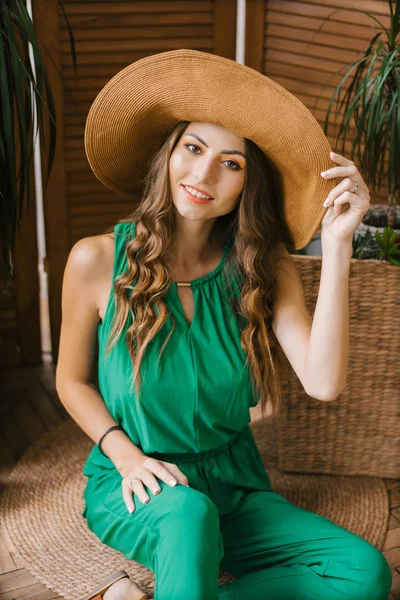 広いつばの帽子をかぶった緑のジャンプスーツで明るい化粧をした美しい笑顔の女の子が座り 帽子に手を握っている — ストック写真