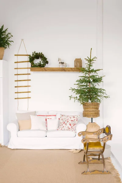 Stylish Scandinavian decor in the living room for Christmas or New Year: a white sofa with pillows, a Christmas tree, a wooden rocking chair, Souvenirs and a Christmas wreath