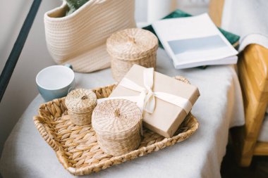 Bej renkli saten kurdeleyle bağlanmış bir hediye kutusu hasır tepside kapaklı saman sepetleri ve bir kupa. Ev konforu